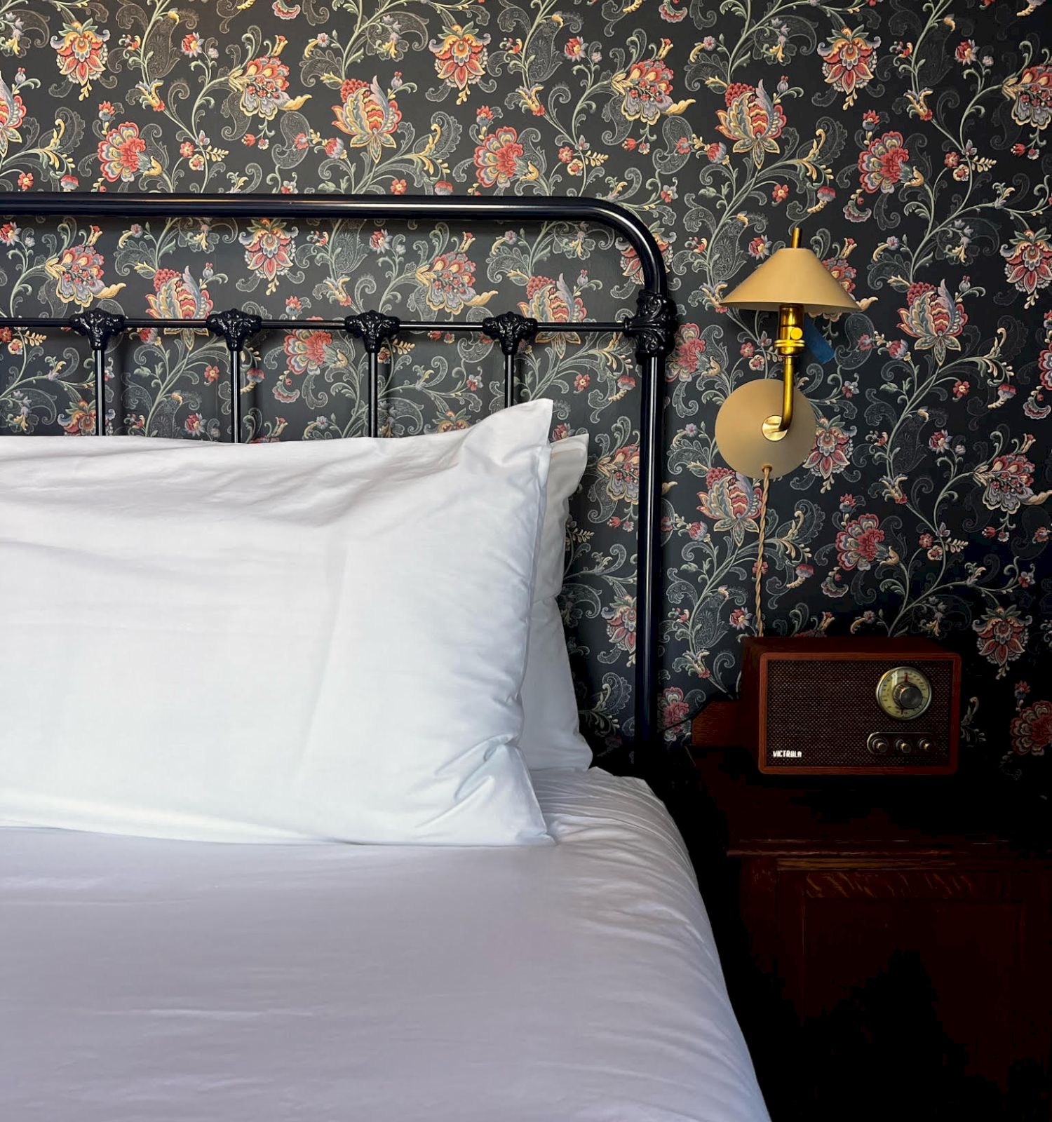 A neatly made bed with white linens, a floral wallpapered wall, a vintage lamp, and a retro radio on a wooden nightstand.