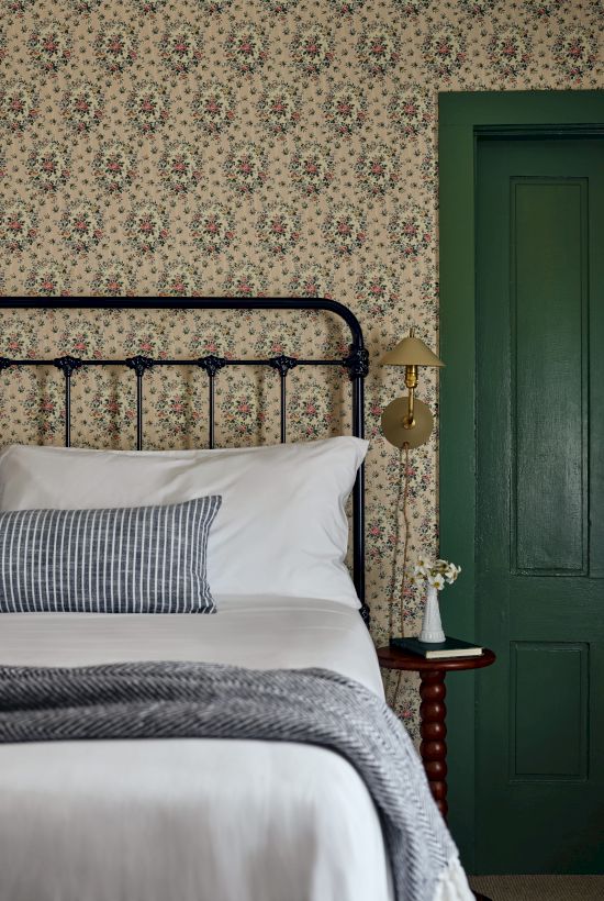 A vintage bedroom features a metal bed with white and striped bedding, floral wallpaper, a green door, and a nightstand with a small decoration.