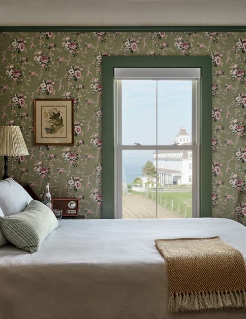 A cozy bedroom with floral wallpaper, a bed with white linens, and a window showing a lighthouse and sea view.