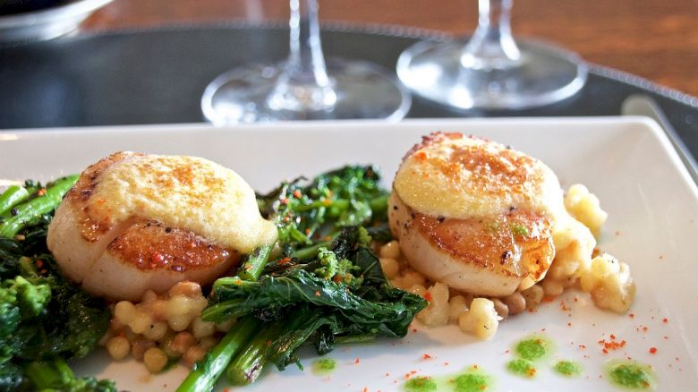 The image shows a plated dish with two seared scallops topped with a creamy sauce, accompanied by sautéed greens and small pasta pearls.