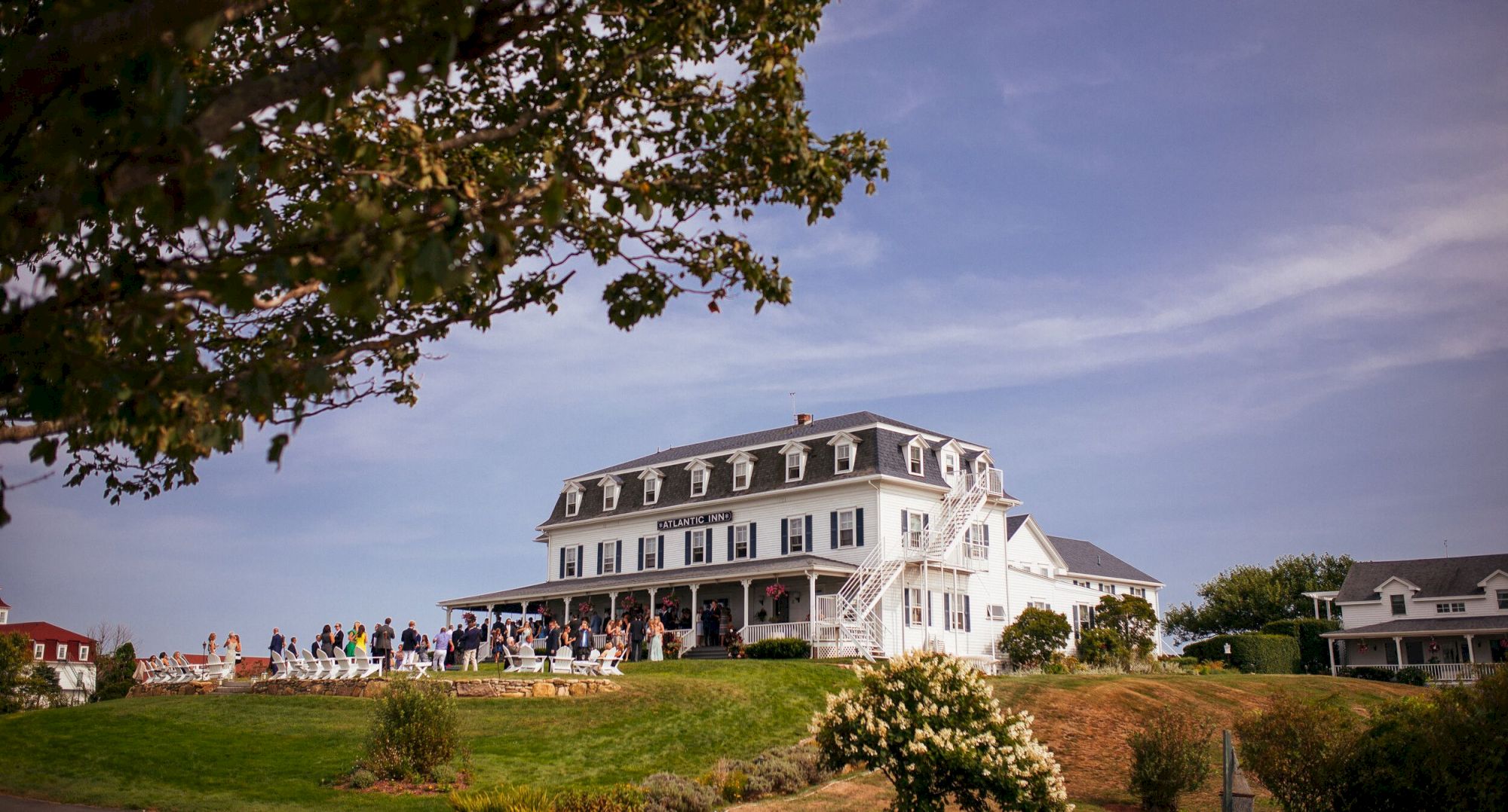 The image shows a large white building with people gathered outside on a grassy hill, under a partly cloudy sky.