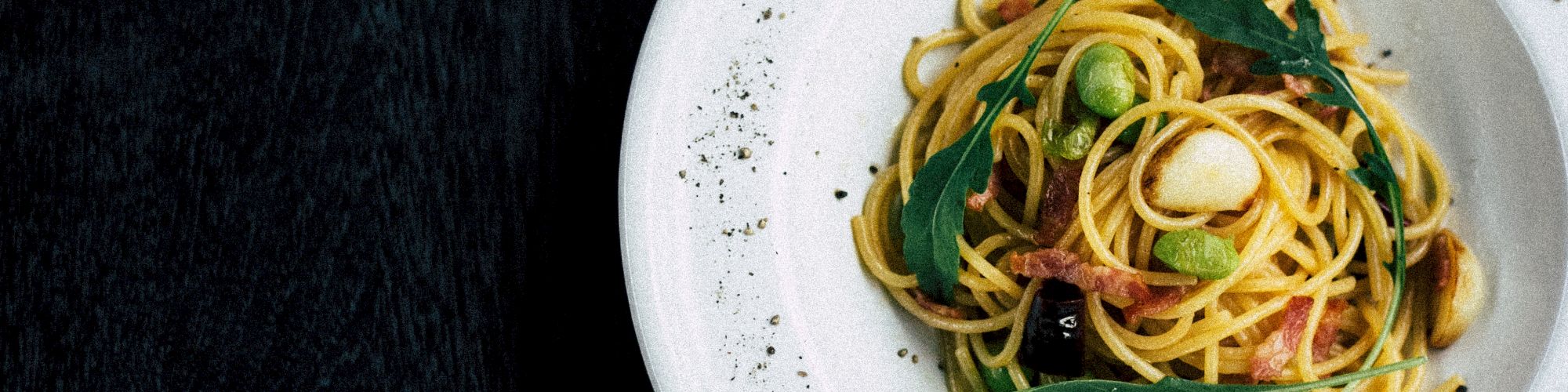 A plate of spaghetti pasta garnished with green herbs, edamame, and possibly a light sauce, on a dark background.