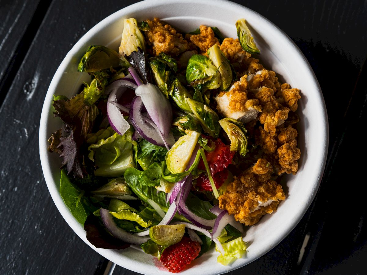 The image shows a bowl containing a salad with mixed greens, red onions, Brussels sprouts, and a portion of what appears to be fried chicken.