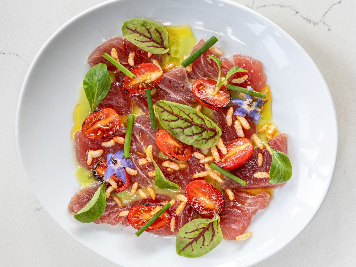 A plate of tuna carpaccio garnished with cherry tomatoes, pine nuts, microgreens, and edible flowers on a white dish in a minimalist setting.