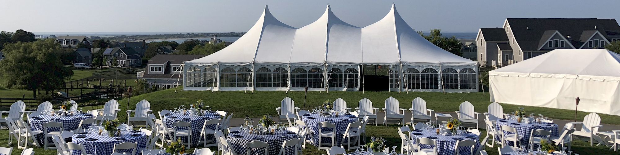 The image shows an outdoor event setup with white tables and chairs arranged on a lawn in front of a large white tented structure.