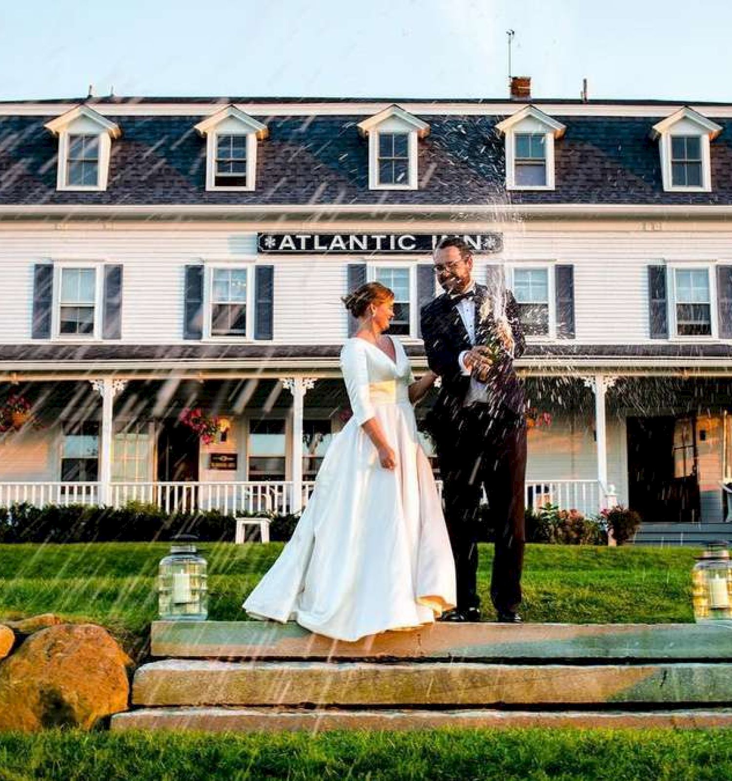 A couple is standing on the steps in front of the Atlantic Inn, with a scenic background and water sprinkling around them.