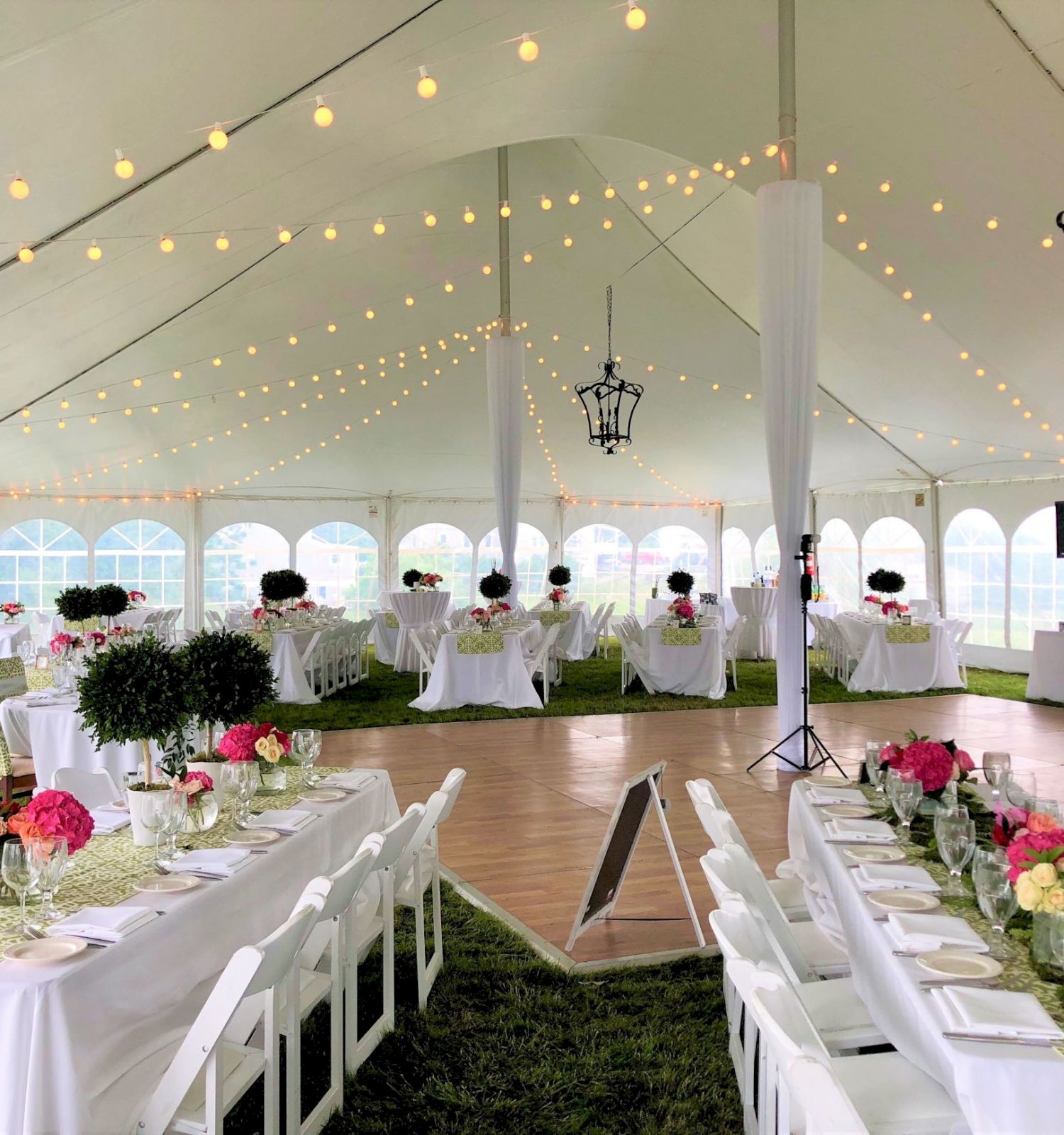 A beautifully decorated event tent with white tables and chairs, floral centerpieces, string lights, and lanterns, likely set up for a wedding or party.