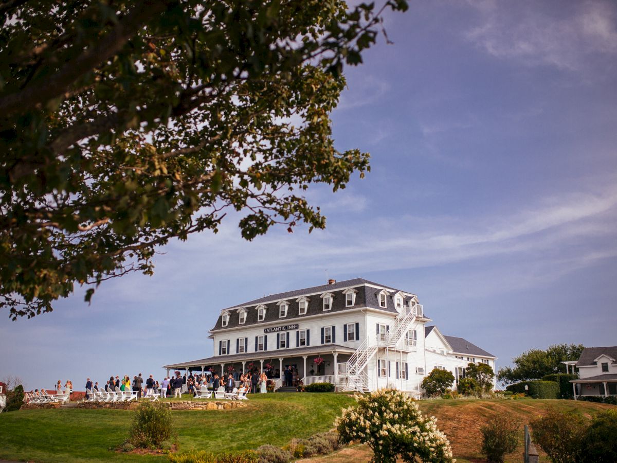 The image shows a large, white building with a slanted roof, possibly a hotel or inn, surrounded by greenery and a clear, blue sky.