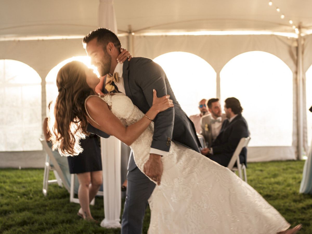 A bride and groom are dancing under a tent with string lights, surrounded by a few seated guests in the background, sun setting.