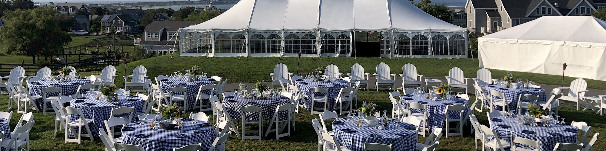 The image shows an outdoor event setup with neatly arranged tables and chairs, in front of a large white tent on a grassy area, with houses in the background.