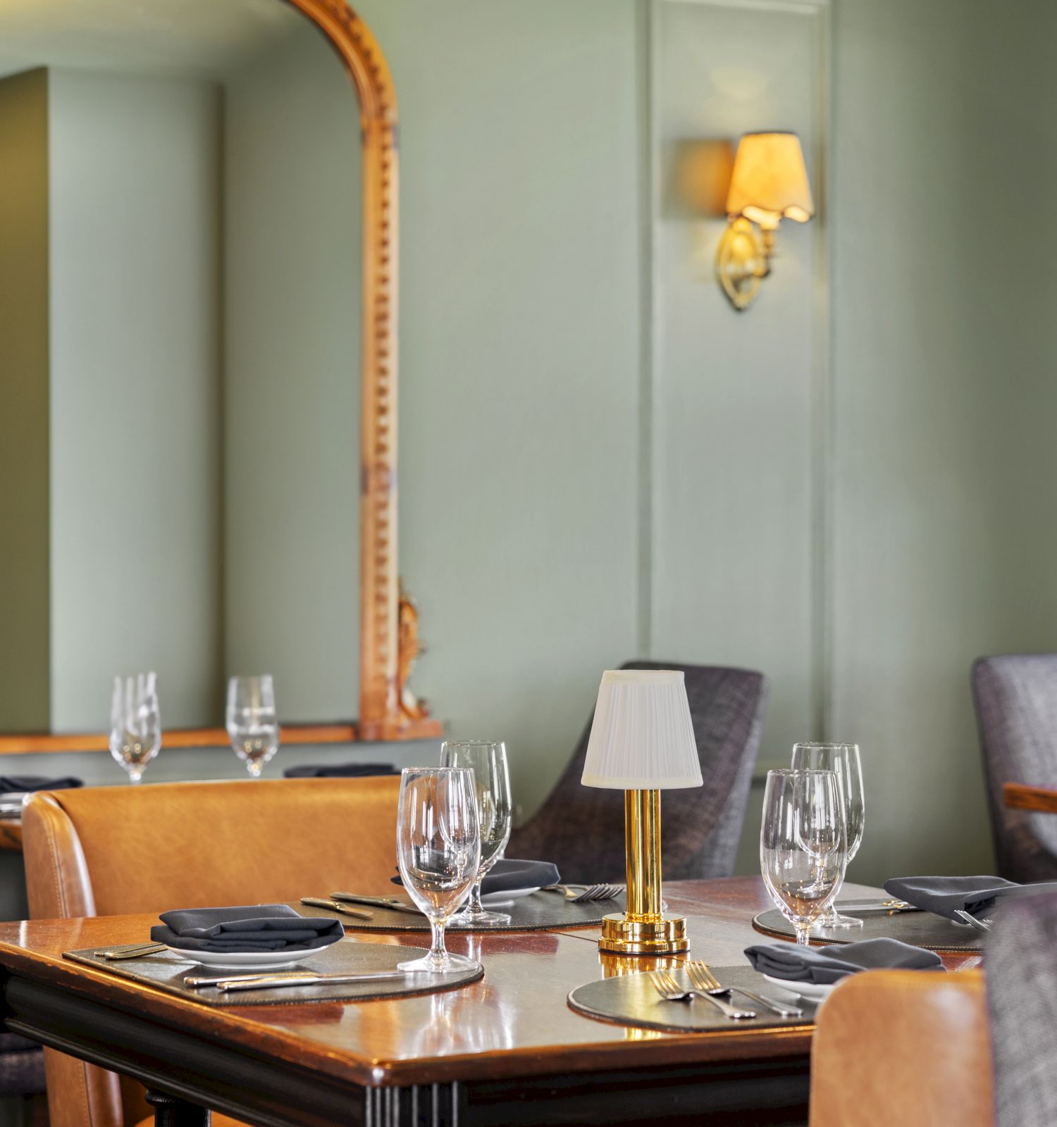 A cozy restaurant setup with tables set for dining, including wine glasses, napkins, a small lamp centerpiece, and a large decorative mirror.