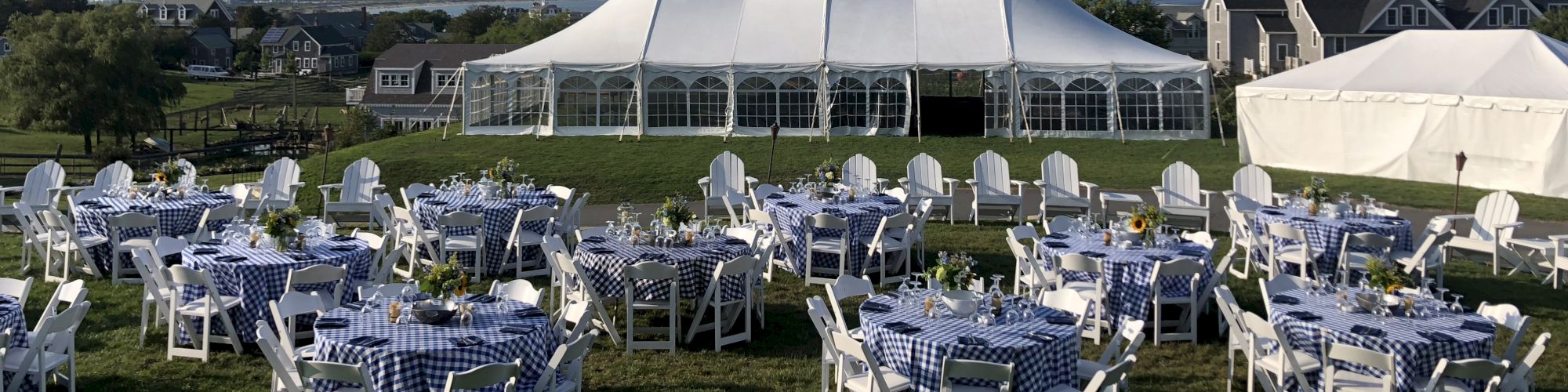 Image showing outdoor event setup with white tents, round tables with checkered cloths, and white chairs on green lawn under clear sky.