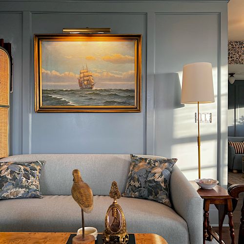 The image shows a cozy living room with a sofa, decorative cushions, a painting of a ship, a lamp, and a coffee table with a book and decor items.