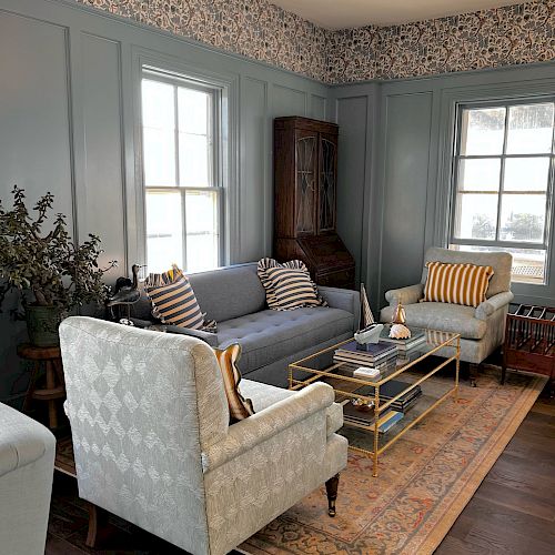 A cozy living room features a sofa, two armchairs, a coffee table, a rug, and a tall wooden cabinet, with blue walls and patterned wallpaper.