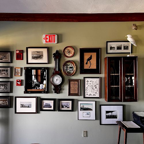 A wall with framed pictures, a clock, and a mirror, adorned with a white bird figurine, cabinets, and an exit sign positioned on top.