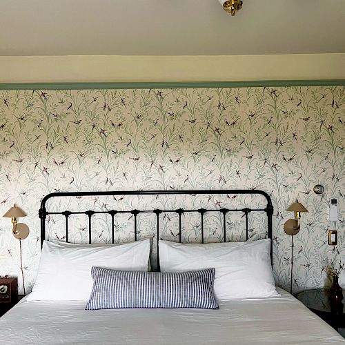 A neatly made bed with white linens and a striped pillow against a floral wallpaper backdrop, flanked by wall-mounted lamps.