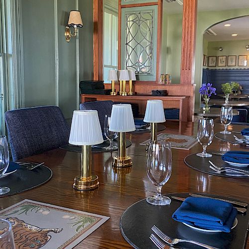 A dining setting with a wooden table, place settings, and small lamps. The background features a mirror, wood details, and softly lit ambiance.