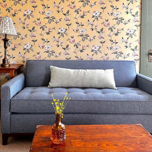 A cozy living room with a gray sofa, a floral pillow, a wooden coffee table, a side table with a lamp, and a floral wallpaper background.