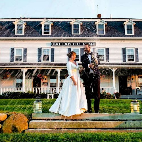 A couple in formal attire stands on steps in front of Atlantic Inn, surrounded by greenery and chairs, with water spraying above them.