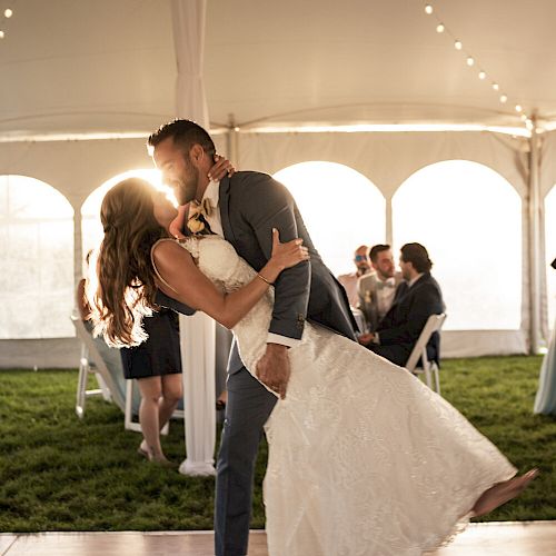 A couple shares a dance under a tent with string lights while guests watch in the background, in what appears to be a wedding reception.