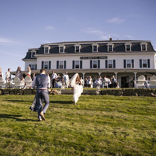 A wedding couple walks towards a large building with a crowd outside, set against a clear blue sky, surrounded by green grass.