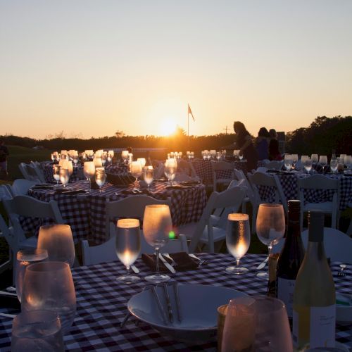 An outdoor dining setup with checkered tablecloths, glassware, and wine bottles, set against a sunset background with people in the distance.