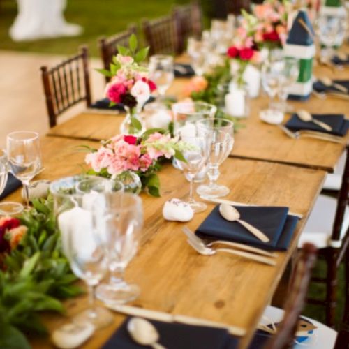 A long wooden table is set for a formal event with floral centerpieces, glassware, utensils, and folded napkins, surrounded by chairs.