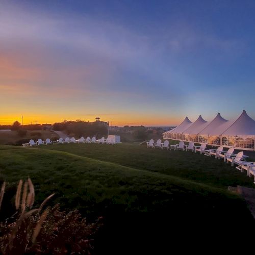 The image showcases a serene outdoor setting at sunset, with white tents and white chairs arranged on a grassy lawn.