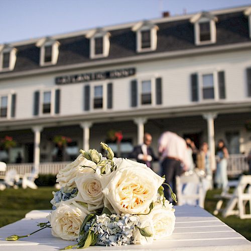 A bouquet of white roses is in focus in the foreground, with a large white building and people in the background.