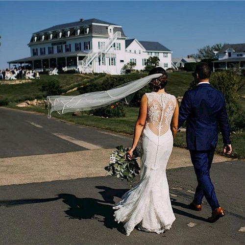 A newlywed couple in wedding attire walks down a path, with a large, elegant building in the background and guests gathered on the lawn.