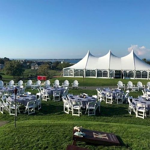 The image shows an outdoor event setup with round tables and white chairs on a grassy field, with a large white tent in the background under a clear sky.