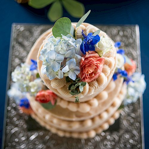 A decorative tiered cake with intricate icing and adorned with colorful flowers, including blue, white, and orange blooms, on a blue tablecloth.