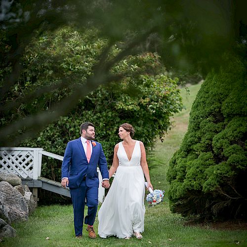 A couple is walking hand in hand outdoors; the woman is in a white dress holding a bouquet, and the man is in a blue suit with a pink tie.