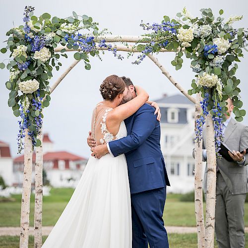 A couple is embracing under a flower-adorned arch outdoors, possibly during a wedding ceremony, with an officiant standing nearby.