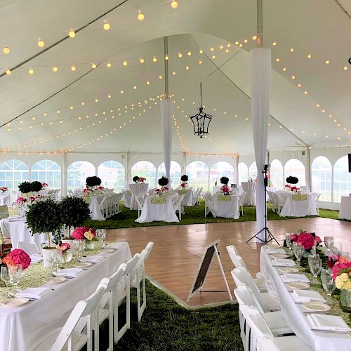 A decorated tent for an event features elegantly set tables with flowers, white chairs, string lights, and a wooden dance floor, indicating a celebration.