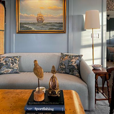 A cozy living room featuring a painting of a ship, a light gray couch with patterned pillows, a decorative table, and a floor lamp.