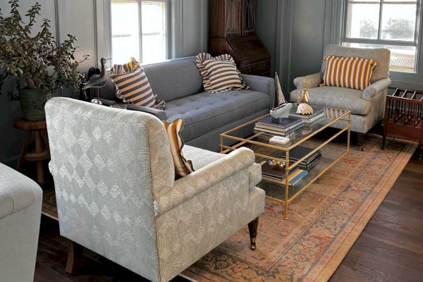 A cozy living room with light blue walls, a gray sofa, two armchairs, a coffee table, a potted plant, and windows with natural lighting, is shown.