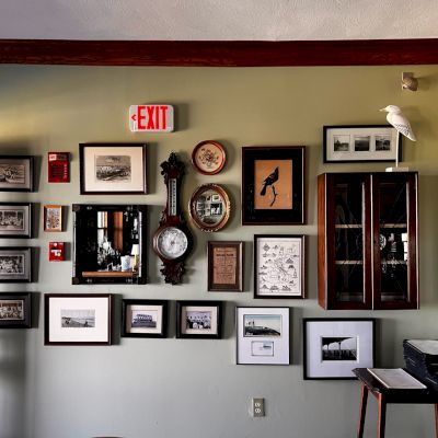 A collection of framed pictures and a clock are mounted on a wall beneath an exit sign, with a white bird figurine on a shelf.