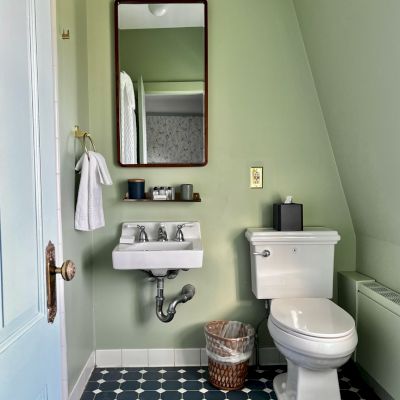 This image shows a small bathroom with a toilet, sink, mirror, towel rack, wastebasket, and green walls. The floor has a patterned design.