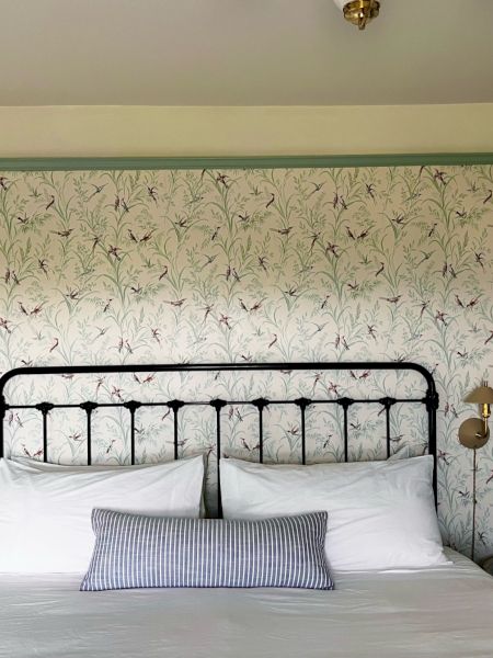 A bedroom with a wrought iron bed, floral wallpaper, two pillows, a striped cushion, two side tables, and wall-mounted lamps on either side.