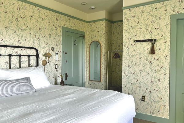 A cozy bedroom with floral wallpaper, a neatly made bed with white linens, a mirror, green door, and wall hooks.