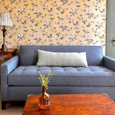 The image shows a cozy living room with a blue sofa, a small wooden table, a floral wallpaper, a lamp on a side table, and a vase with flowers.