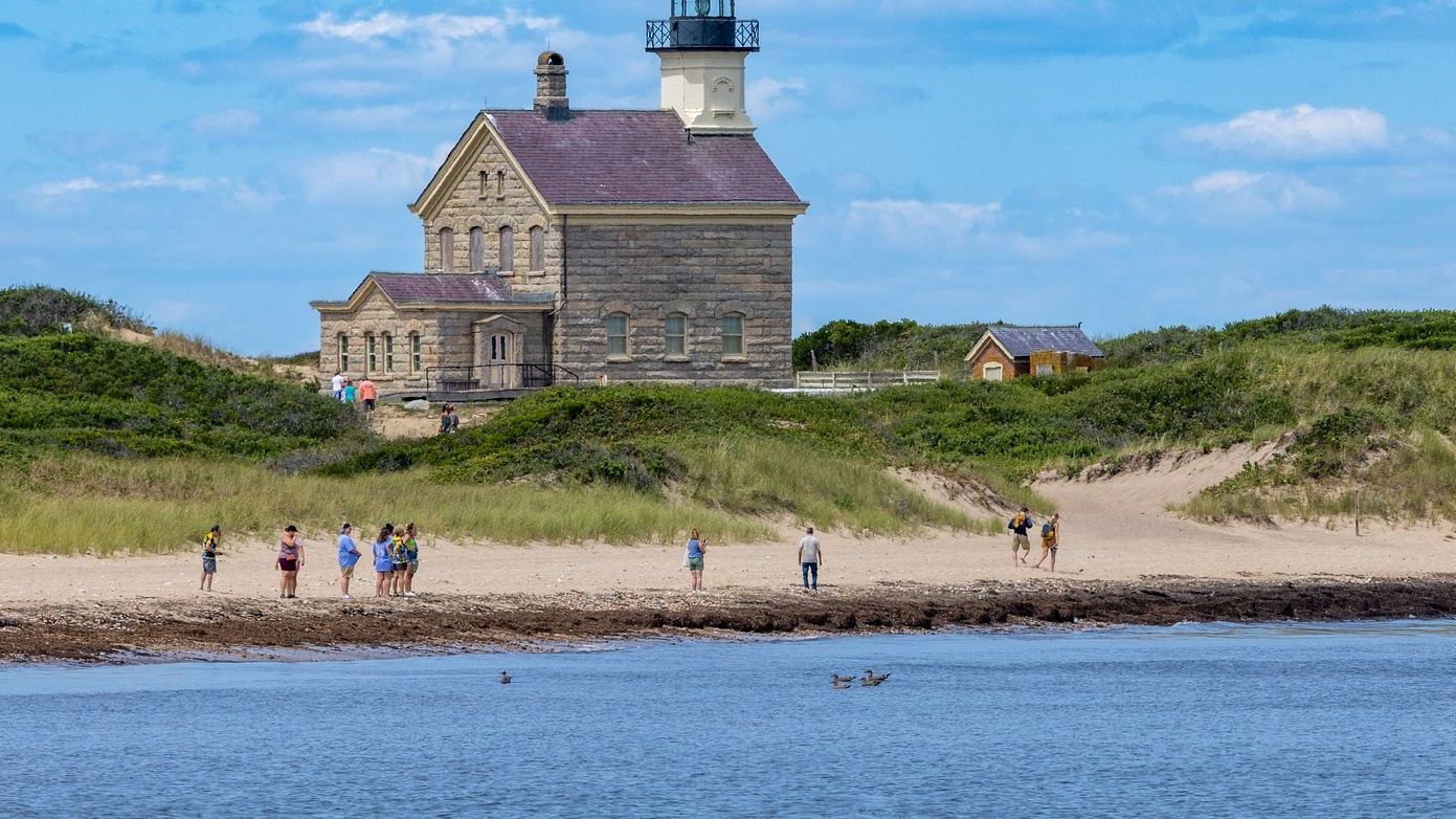 A group of people enjoy a beach near a historic lighthouse situated among grassy dunes with a clear blue sky in the background.