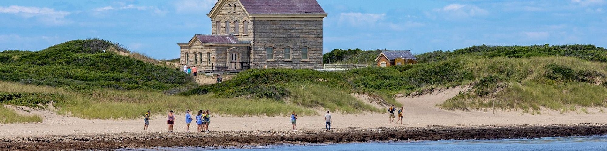 A group of people enjoy a beach near a historic lighthouse situated among grassy dunes with a clear blue sky in the background.