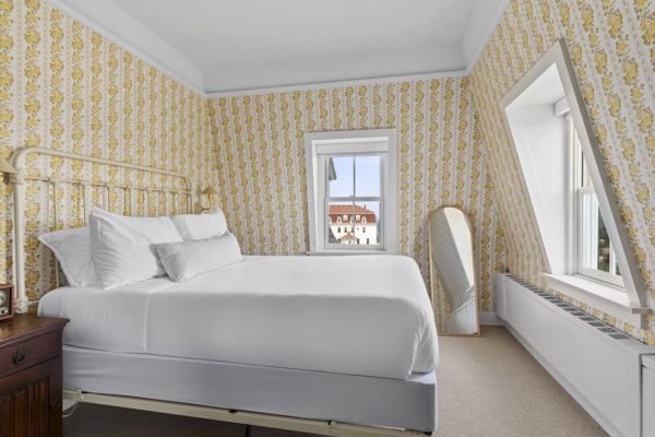 A cozy bedroom with floral wallpaper, a double bed with white linens, a vintage bedside table, a standing mirror, and a window with a view.