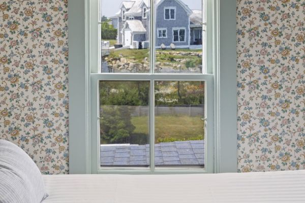 The image shows a window view from a room with floral wallpaper, looking out to a house with a large yard and some rocks in the background.