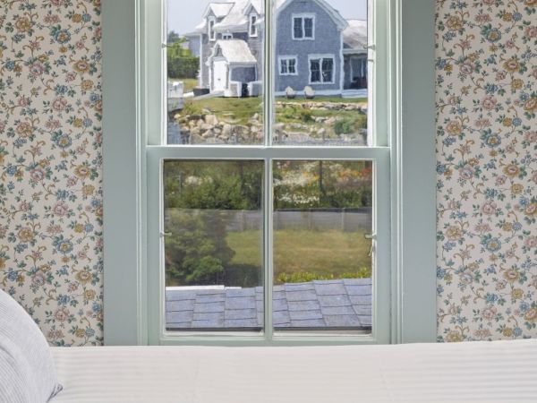 The image shows a window view from a room with floral wallpaper, looking out to a house with a large yard and some rocks in the background.