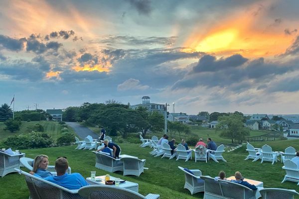 People are sitting on outdoor furniture, enjoying a sunset view over a scenic landscape with scattered houses in the background.