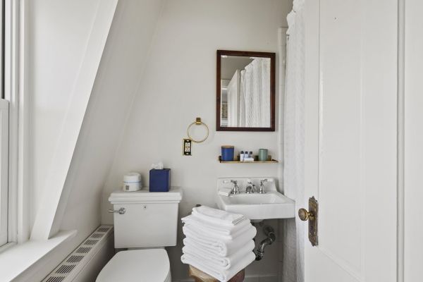 A small, clean bathroom with a toilet, sink, mirror, and stacked white towels. It includes a window and some toiletries on a wall-mounted shelf.