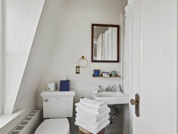 A small, clean bathroom with a toilet, sink, mirror, and stacked white towels. It includes a window and some toiletries on a wall-mounted shelf.